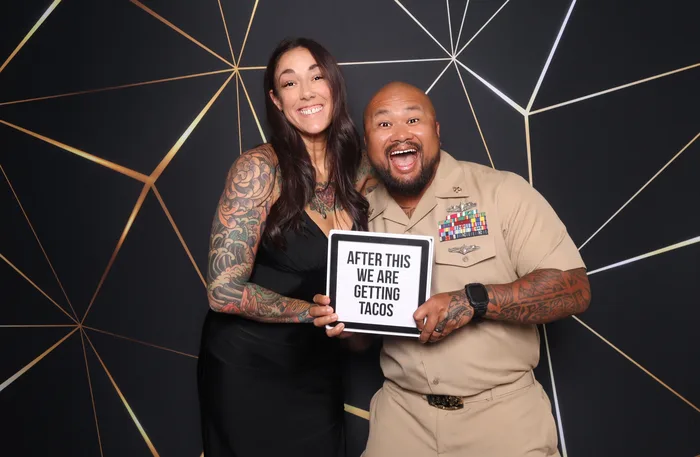 A sailor in Navy khakis posing with his date and an 'After This We Are Getting Tacos' prop sign in front of a black and gold geometric backdrop — photo booth at a military event in Virginia Beach