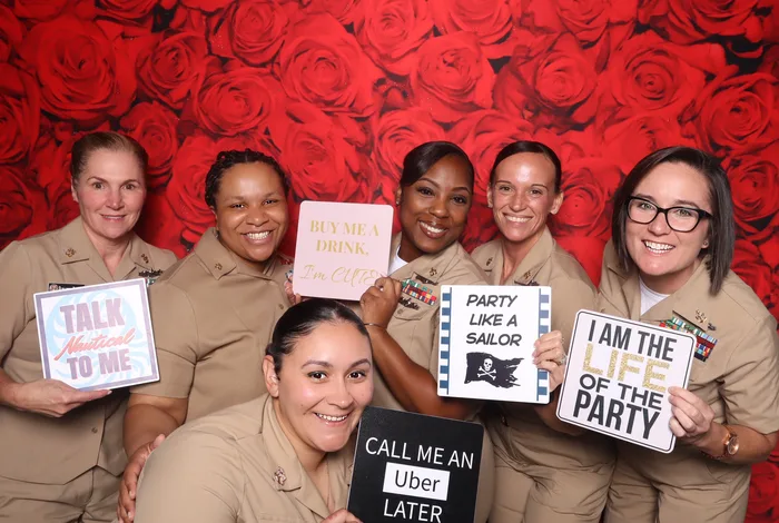 Six women sailors in Navy khaki uniforms holding playful photo booth prop signs in front of a red roses backdrop at a military event in Virginia Beach