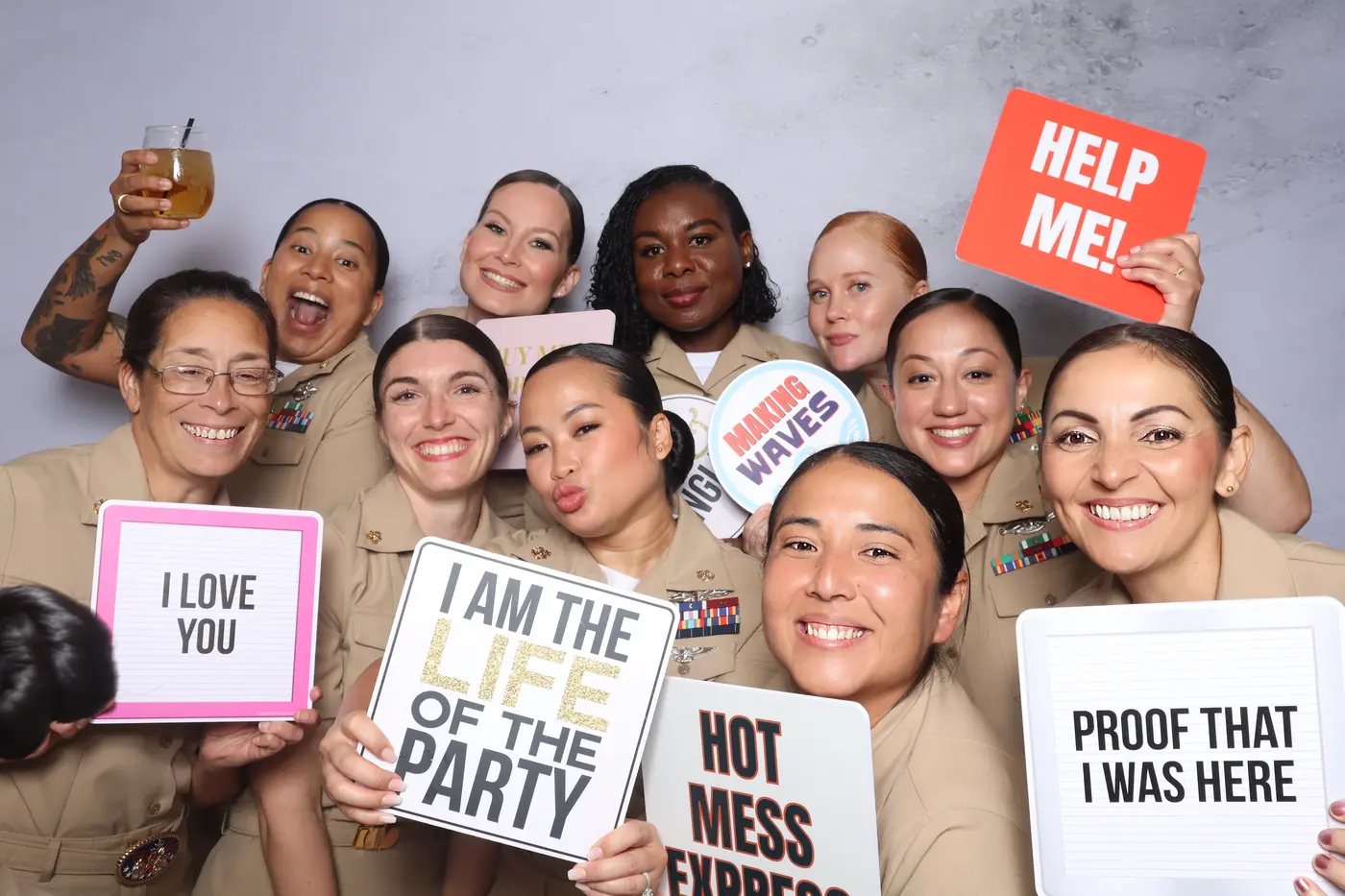 Group of sailors in khaki uniforms laughing with fun prop signs at a Khaki Ball photo booth, Hampton Roads
